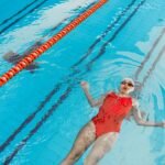 A woman in a red swimsuit floats on her back in an indoor swimming pool, enjoying a relaxing swim.
